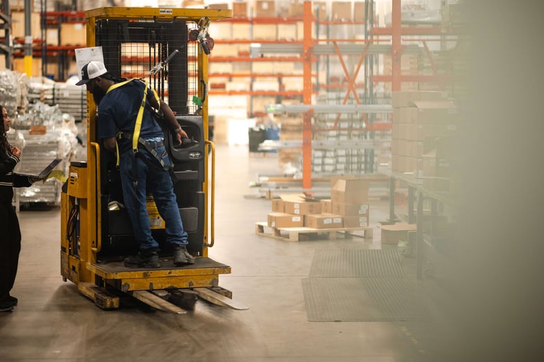 A warehouse worker in a safety harness operates a yellow lift truck while speaking with a colleague holding a laptop. They are surrounded by boxes, shelving, and industrial inventory inside a well-lit distribution center